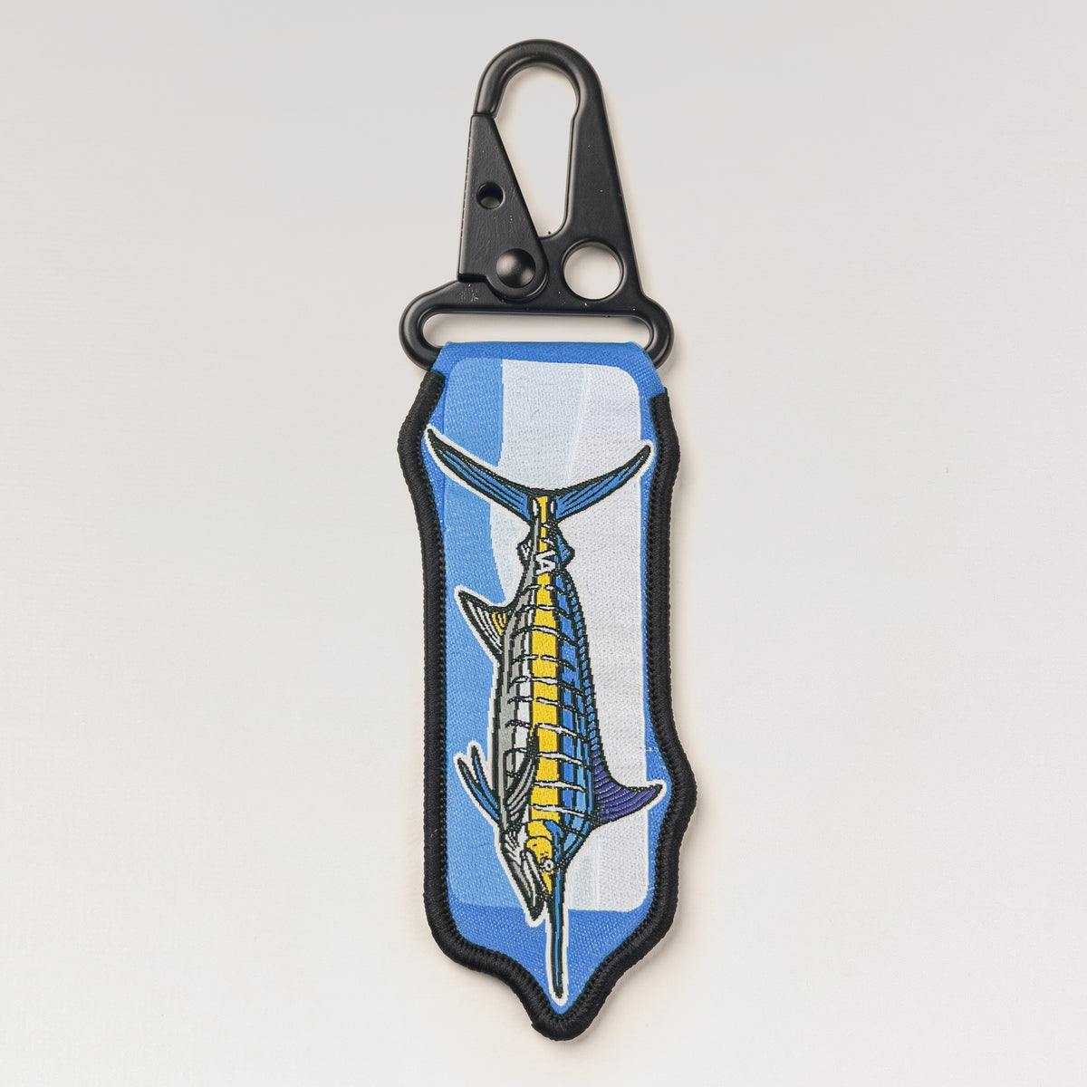 Blue Marlin woven keychain, jacquard fabric tag with black metal carabiner clip, on a light studio background