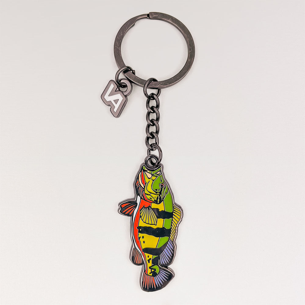 Peacock Bass keychain, black nickel hard enamel charm with the VEDUTA DESIGN VA logo charm and chain, on a light studio background