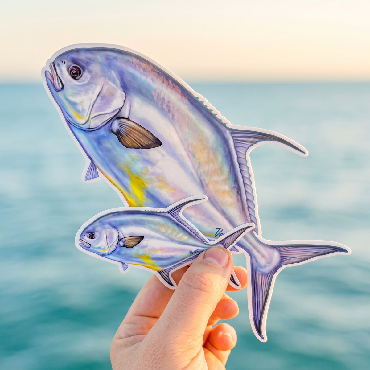 Permit stickers in two sizes held in hand with the ocean at sunset behind